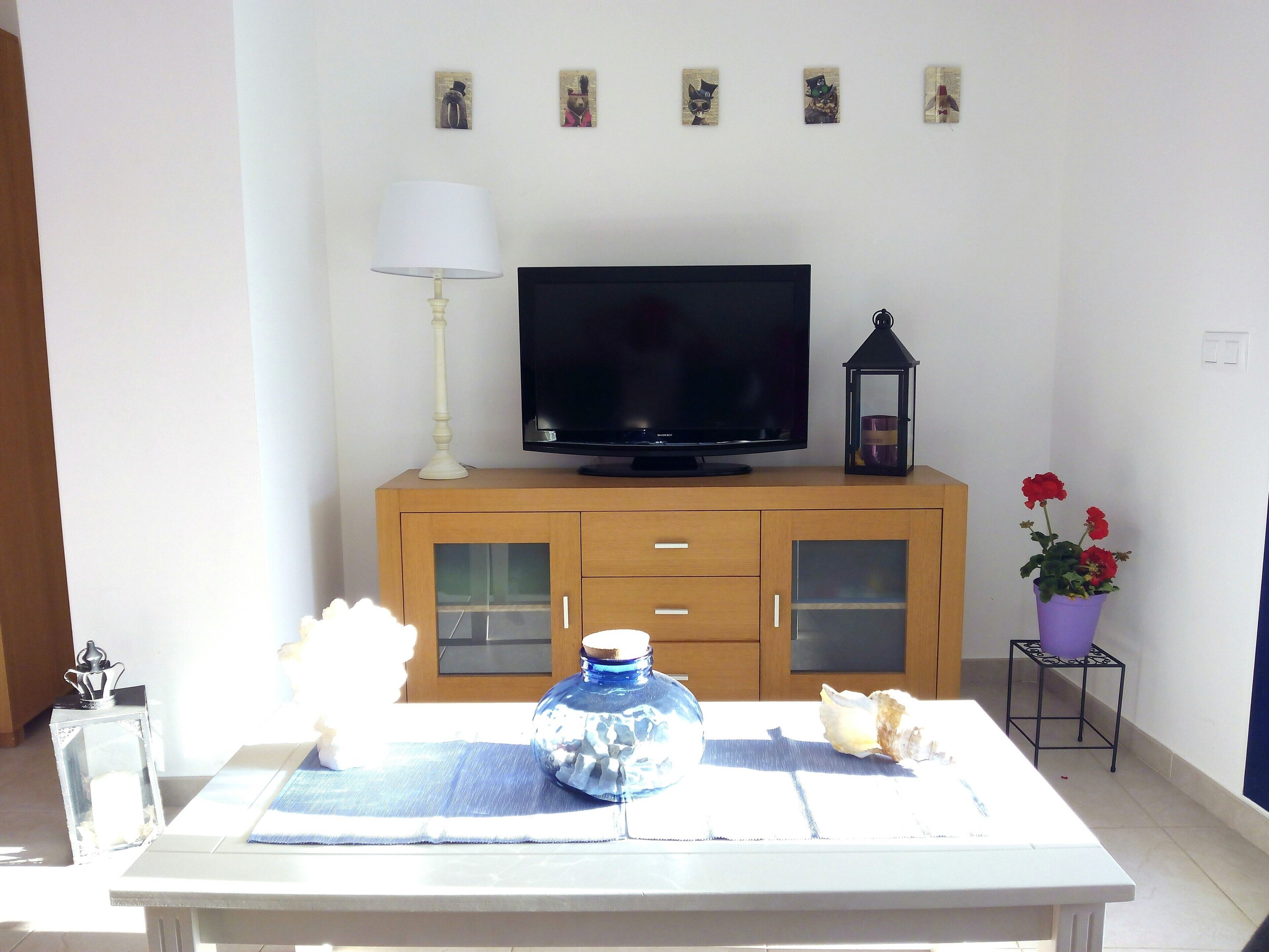 Una sala de estar con un televisor sobre un mueble de madera, una mesa con un florero y flores, y una lámpara.