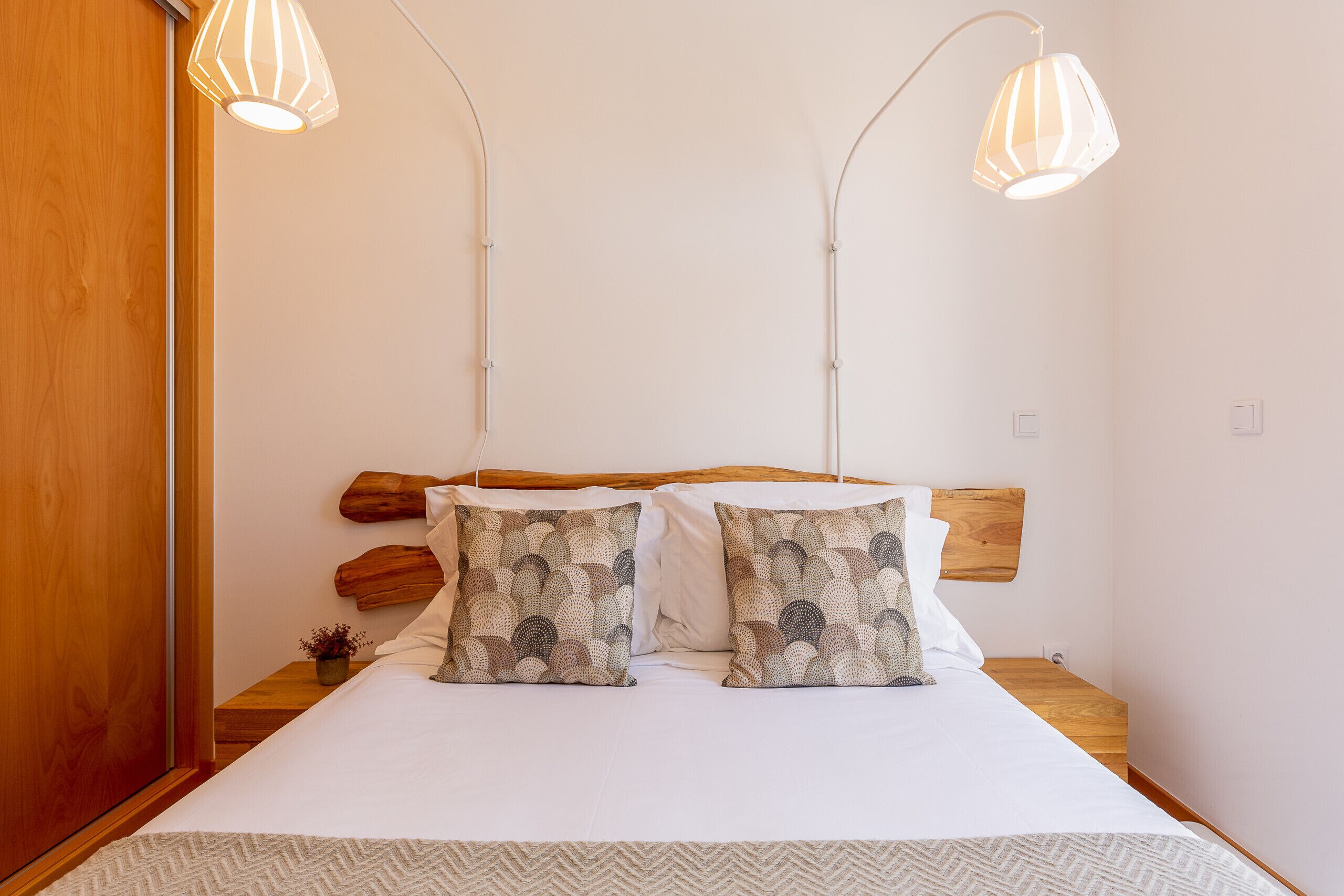 A neatly made bed with two patterned pillows, a wooden headboard, and two pendant lights.