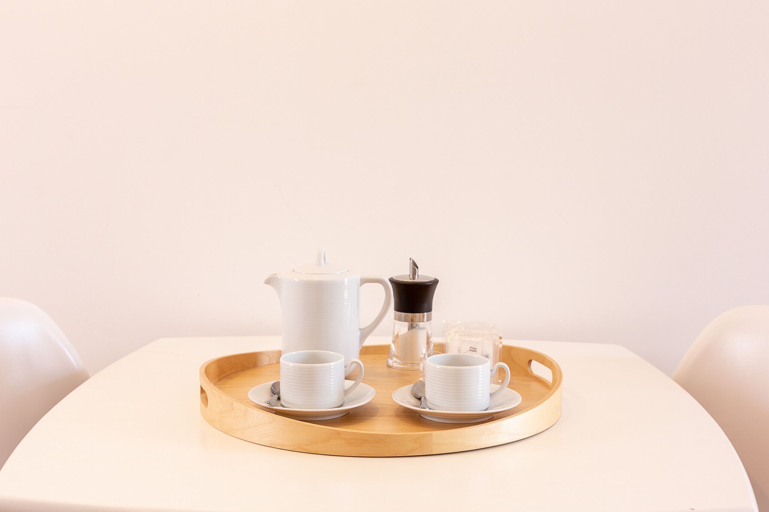 A white table with a tray holding a white coffee pot, two white cups with saucers, and a small glass container.