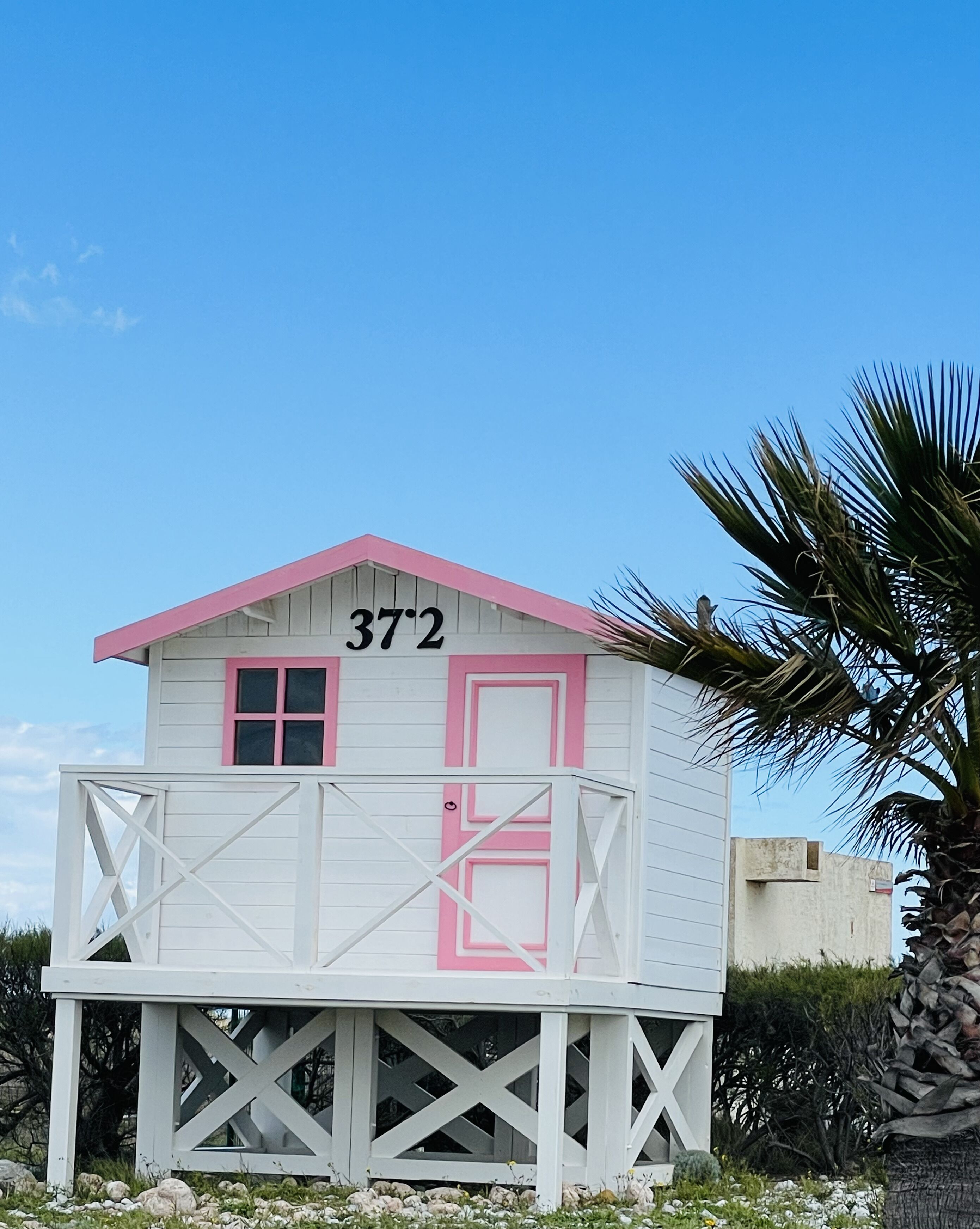A small, single-story building with a pink door and window, marked with the number 372, featuring a balcony with a white railing.