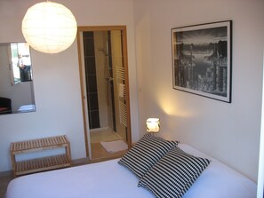 Een hotelkamer met een bed, een houten bankje, een douche, een kunstwerk van een stadsgezicht in een lijst en een hangende lamp.
