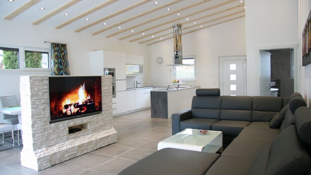 Een moderne woonkamer met een flatscreen-tv, een stenen openhaard, een grijze bank en een keuken op de achtergrond.
