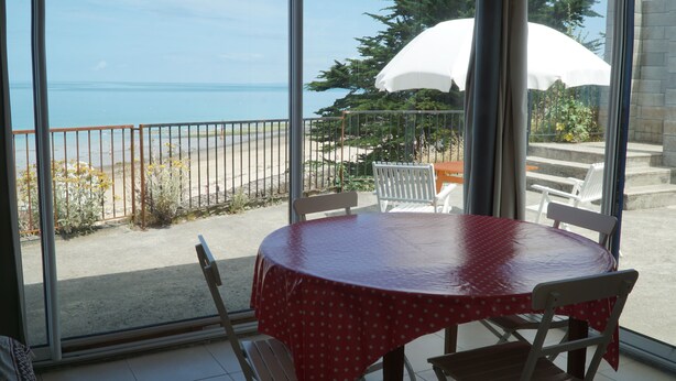 A round table with chairs, a white umbrella, and a view of the sea through large windows.