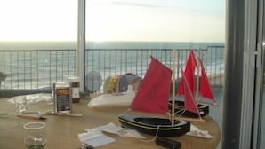 A table with a model sailboat, a glass, and some papers on a balcony overlooking the sea.