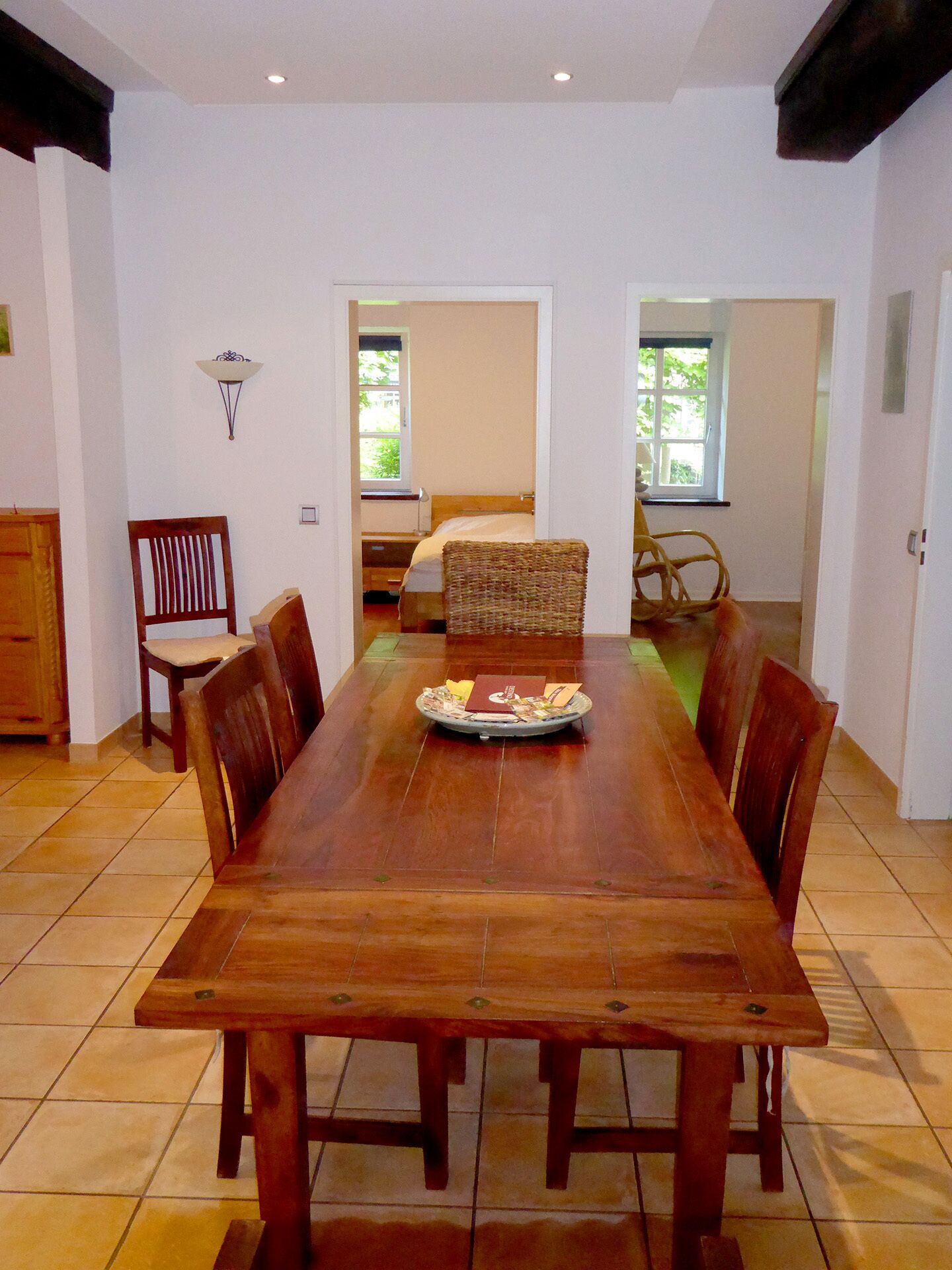 Een ruime eethoek met een lange houten tafel, omringd door stoelen, een bijzettafeltje met een dienblad en uitzicht op een slaapkamer door een open deur.