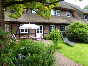 Een traditioneel huis met rieten dak, bakstenen gevel en een tuin met een patio tafel, stoelen en een ligbed.