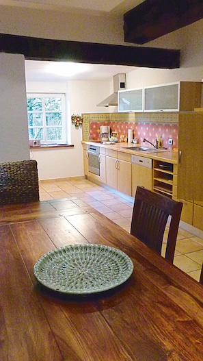 Een eetgedeelte met een houten tafel en stoelen, een keuken met kasten en apparatuur, en een raam met uitzicht op bomen.