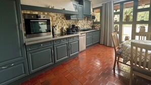 Een keuken met grijze kastjes, een betegelde achterwand, een inbouwoven en een raam met uitzicht op een tuin.
