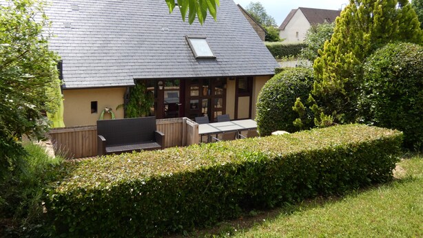 Een huis met een schuin dak, een zichtbare schoorsteen, een houten terras en een goed onderhouden haag.