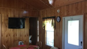 Un interior de madera con una mesa de comedor, sillas, un ventilador de techo, un televisor montado en la pared, una puerta con ventana y un bote de basura.