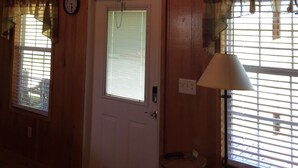 Interior de una cabaña de madera con una puerta, dos ventanas con cortinas, un reloj y una lámpara.
