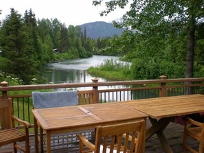 Terraza de madera con mesa y sillas que da a un río, rodeada de vegetación y montañas.