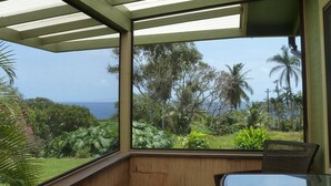 A covered outdoor area with a table and chairs, overlooking a lush landscape with trees and a view of the ocean.