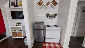 A compact kitchen with a refrigerator, stove, and microwave. There are potted plants on the wall, a small shelf with books, and a red and white checkered rug on the floor. - Dream holiday at a low price: huge travel discounts now available! (Fort Myers)
