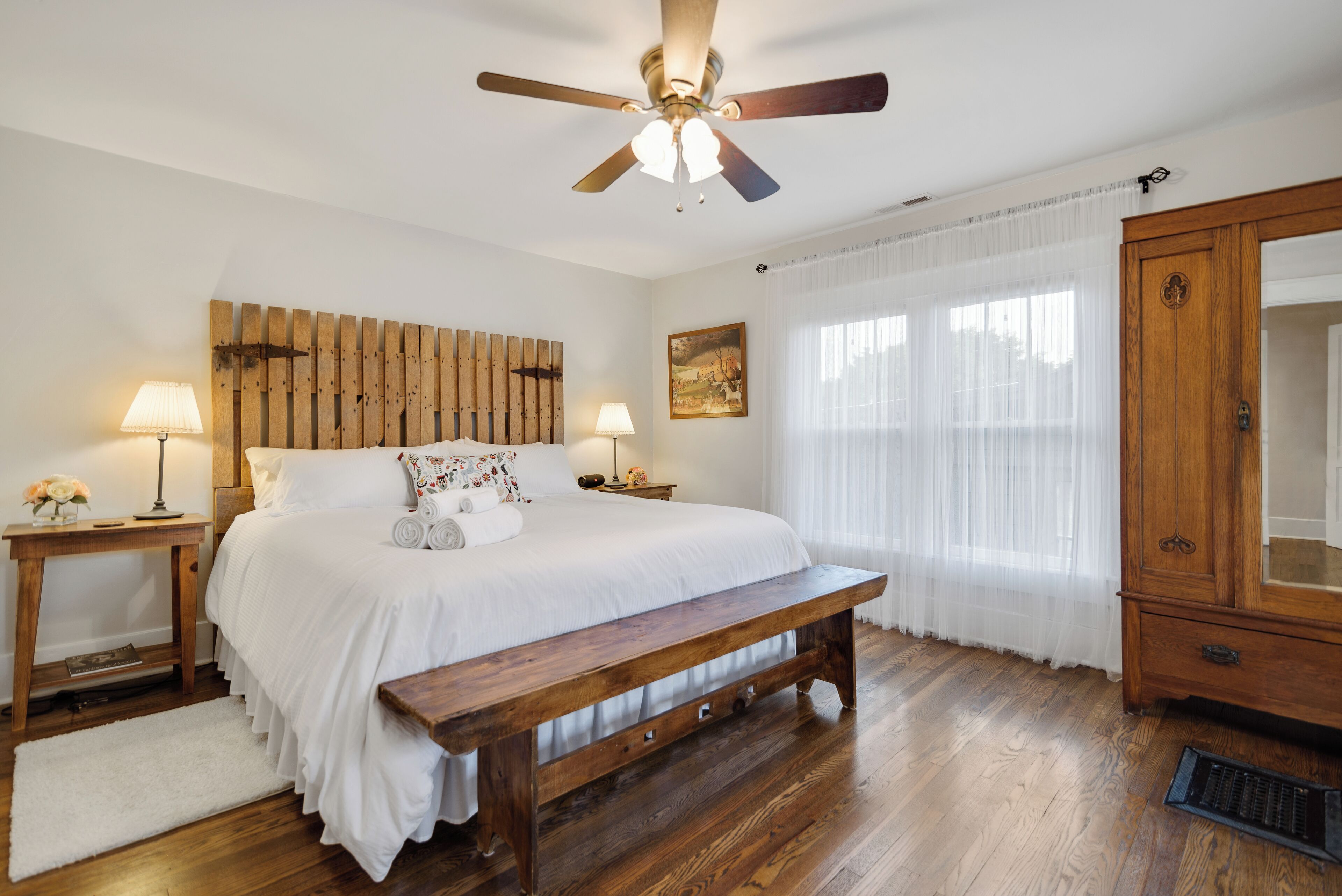 A bedroom with a wooden headboard, a wooden bench, a ceiling fan, a bedside table with a lamp, a bedside table with a lamp, a wooden wardrobe, and a window with sheer curtains.