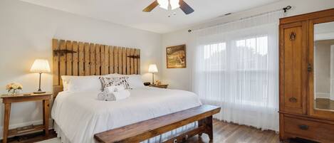 A bedroom with a wooden headboard, a wooden bench, a ceiling fan, a bedside table with a lamp, a bedside table with a lamp, a wooden wardrobe, and a window with sheer curtains.