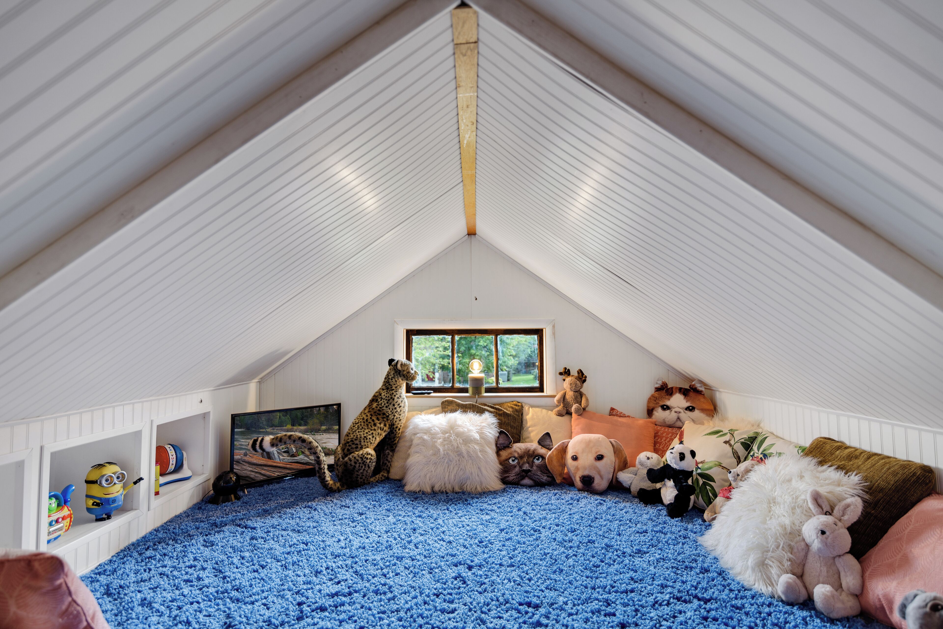 An attic room with a sloped ceiling, a window with a view of greenery, and a blue carpet.