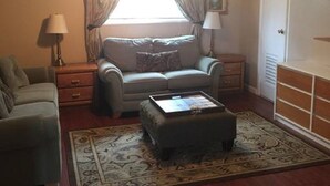 Una sala de estar con sofá, reposapiés y una mesa de centro. Hay un ventilador de techo, una ventana con cortinas y una lámpara.