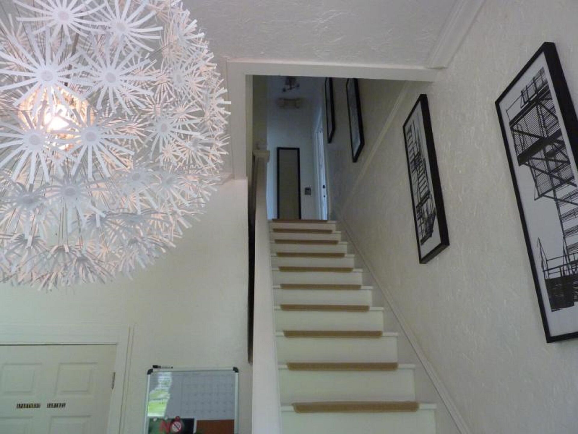 A staircase with white steps and a white wall. There are framed pictures on the wall and a white door at the bottom.