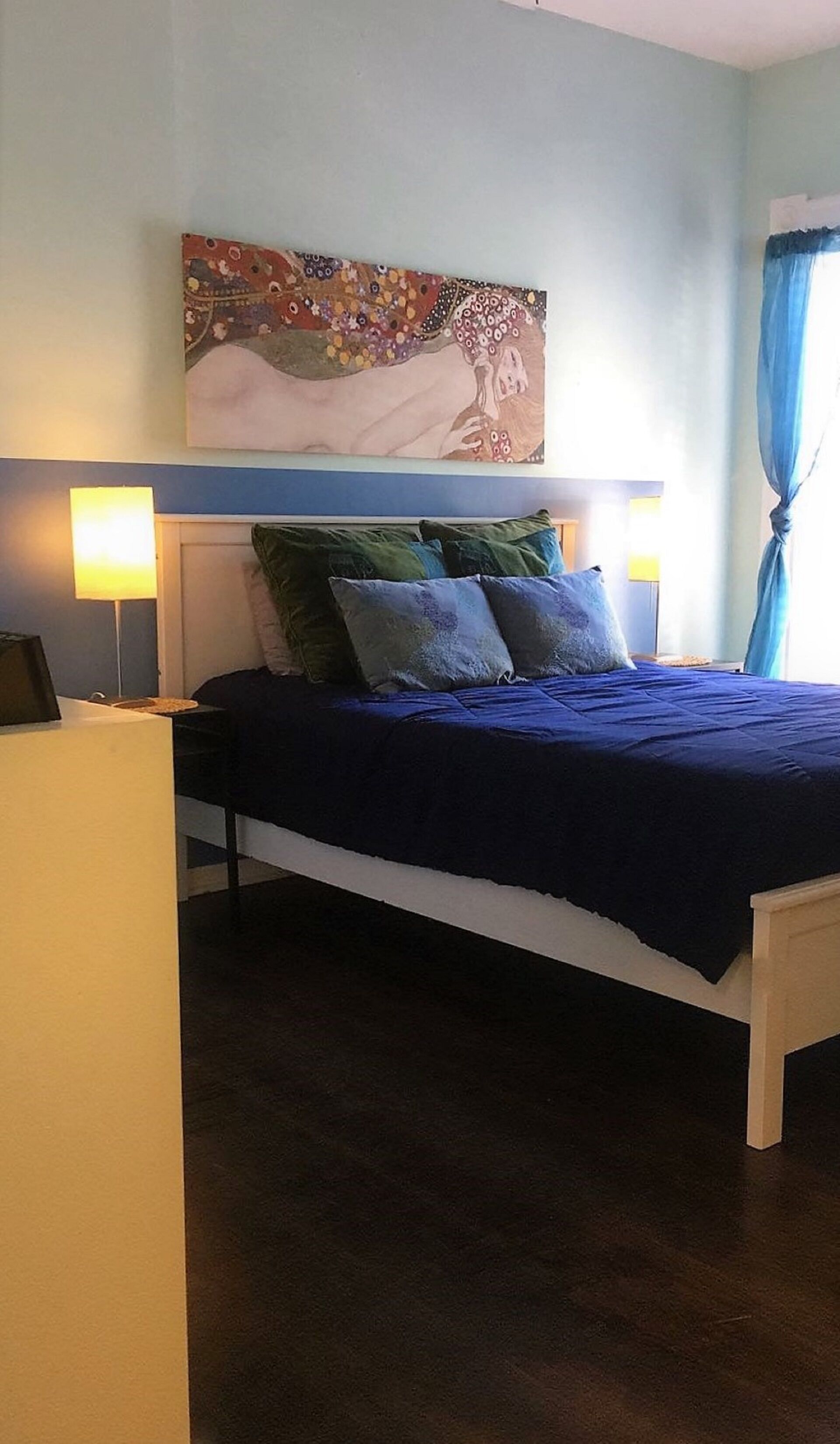A bedroom with a bed, pillows, a nightstand, a lamp, and a painting on the wall.