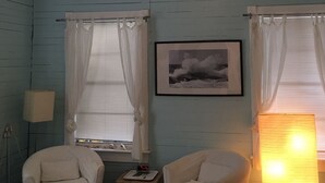 Una habitación con dos sillones blancos, una mesita con un reloj, una fotografía enmarcada en la pared y una lámpara de pie.