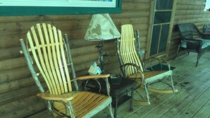 A wooden rocking chair and a chair with a lamp on a porch with wooden flooring.