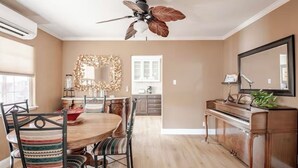 Un coin repas avec une table ronde, des chaises et un ventilateur de plafond. Un piano et un miroir se trouvent sur le côté droit.