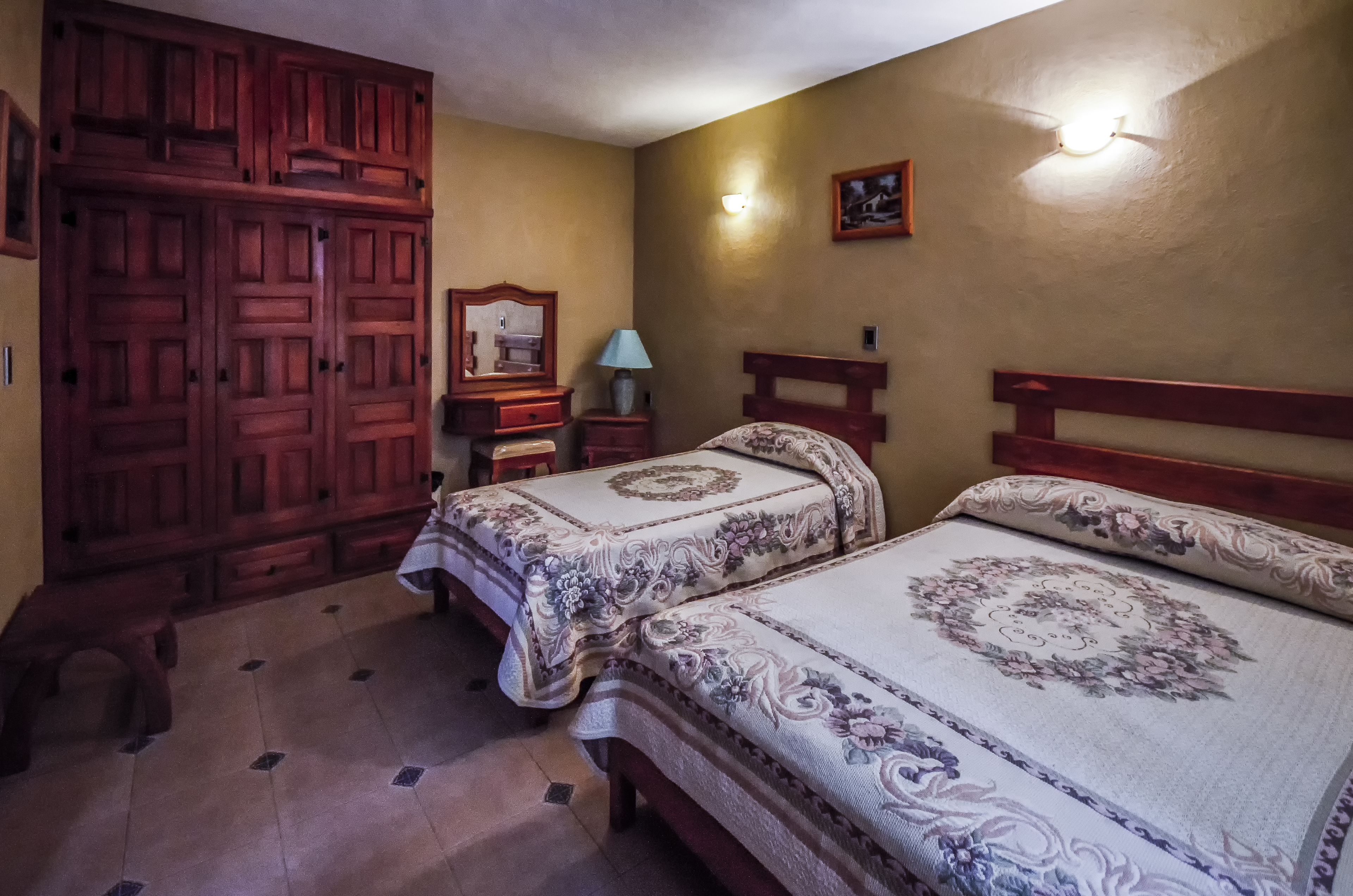 Un dormitorio con dos camas, armario de madera, mesitas de noche, lámpara y aplique de pared.