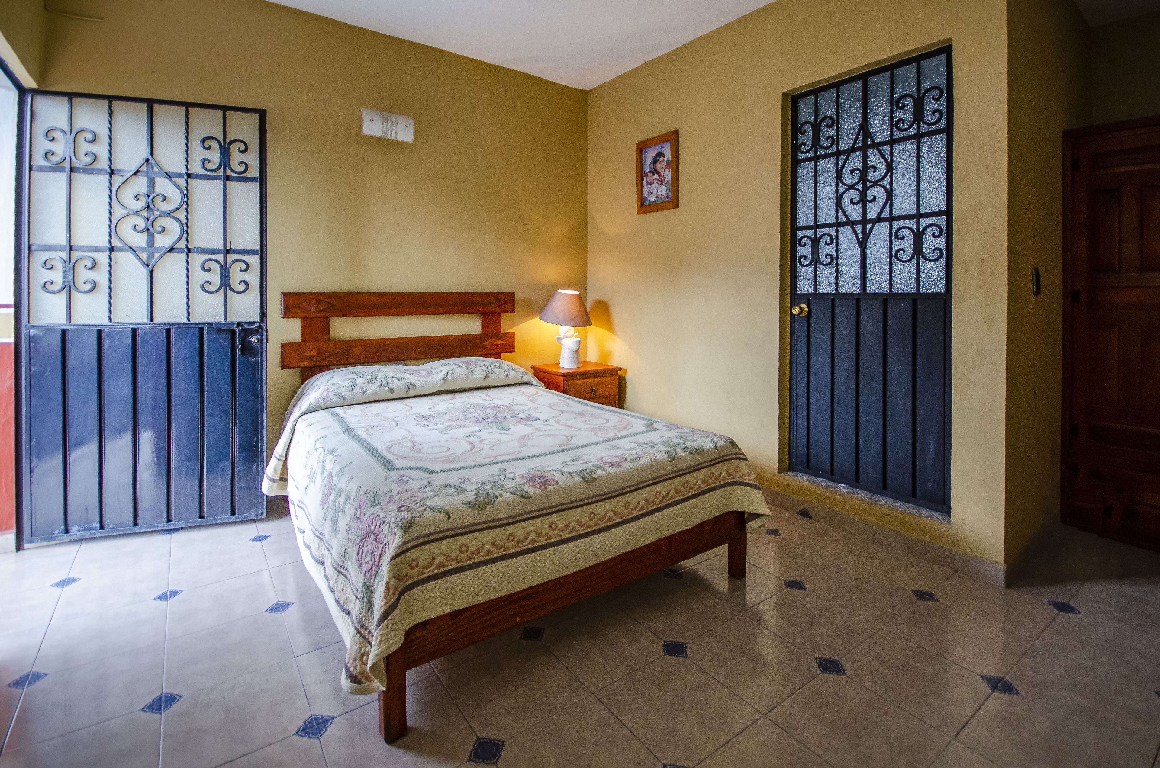 A bedroom with a bed, a nightstand, a lamp, a picture on the wall, and a door with a metal gate.