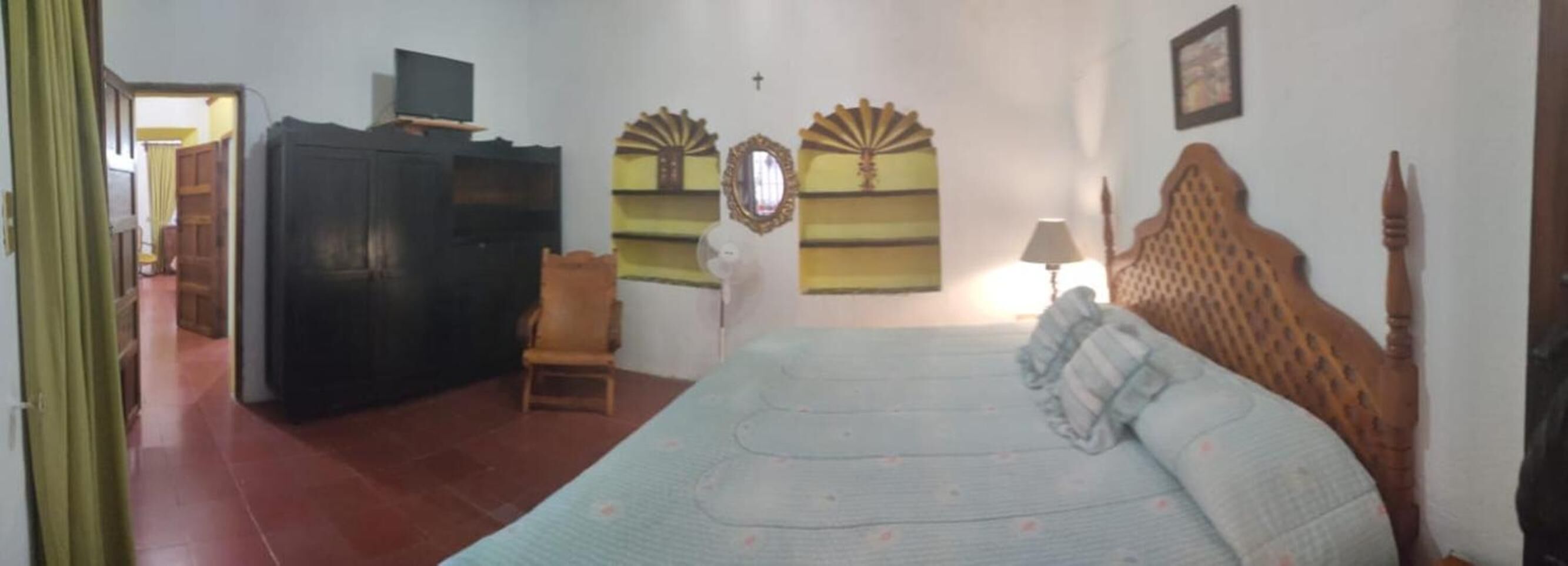 Un dormitorio con cama, cabecera de madera, mesita de noche, una silla, televisor sobre un mueble bajo y espejos decorativos en la pared.