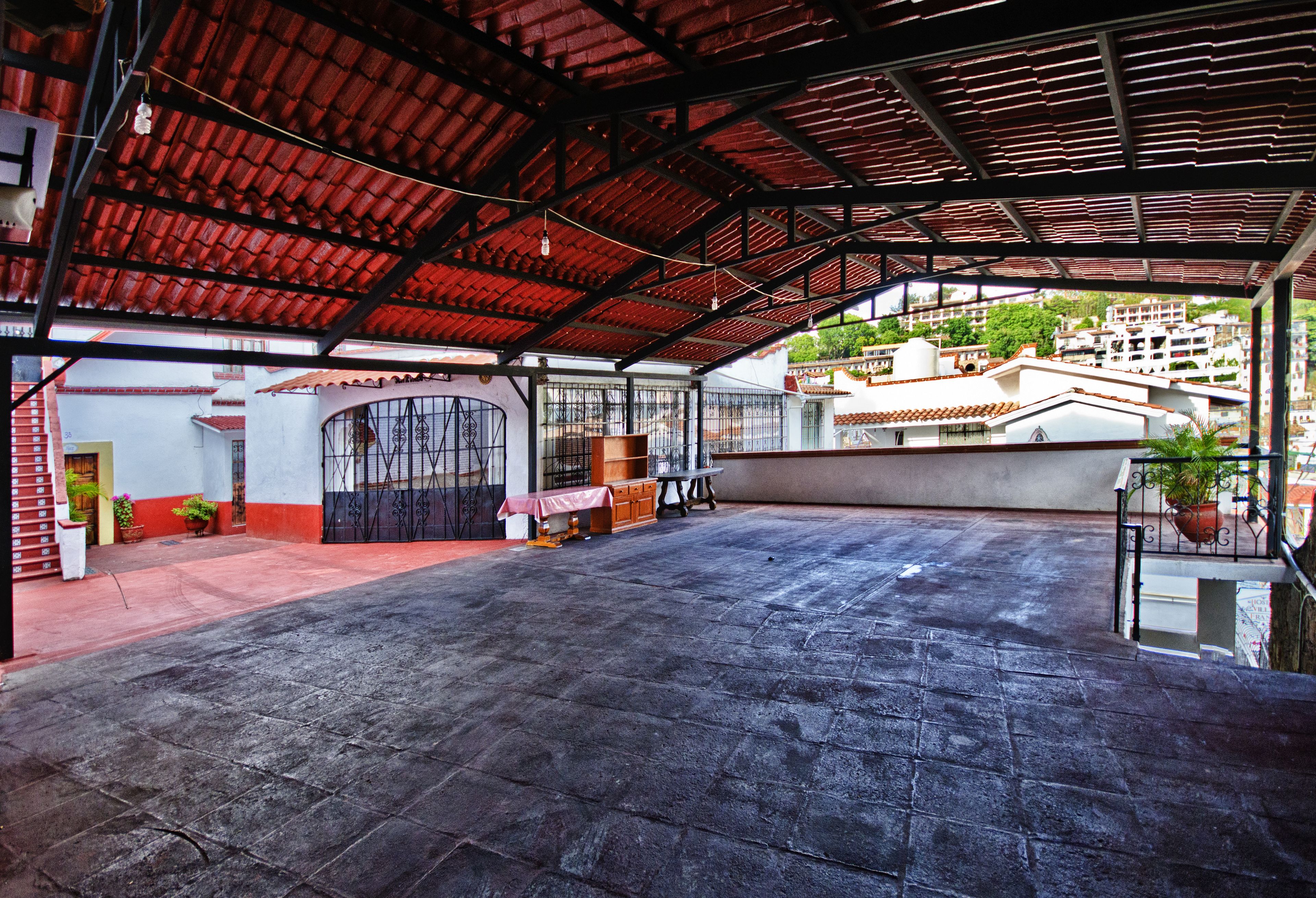 Un patio abierto con suelo de baldosas, una sección cubierta con techo rojo y vistas a edificios residenciales al fondo.