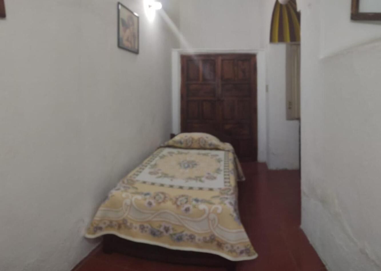 A small, narrow room with a bed, a wooden door, and a light fixture on the wall.