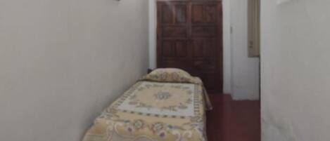 A small, narrow room with a bed, a wooden door, and a light fixture on the wall.