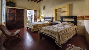 Una habitación con dos camas, una silla de madera, una mesita con una lámpara y un estante.