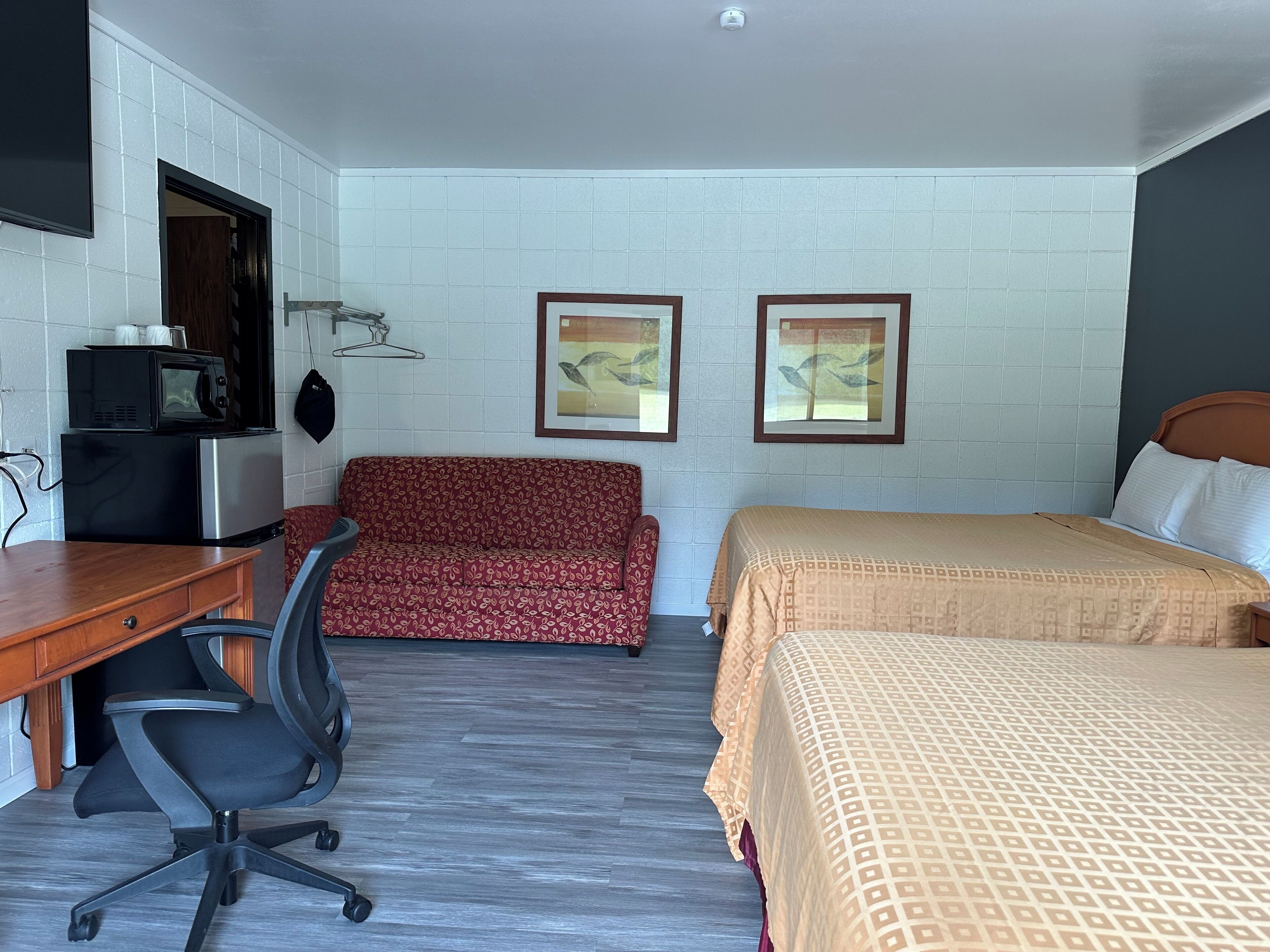 Habitación de hotel con un escritorio de madera, un sofá rojo estampado, una cama con manta color beige y dos cuadros en la pared.