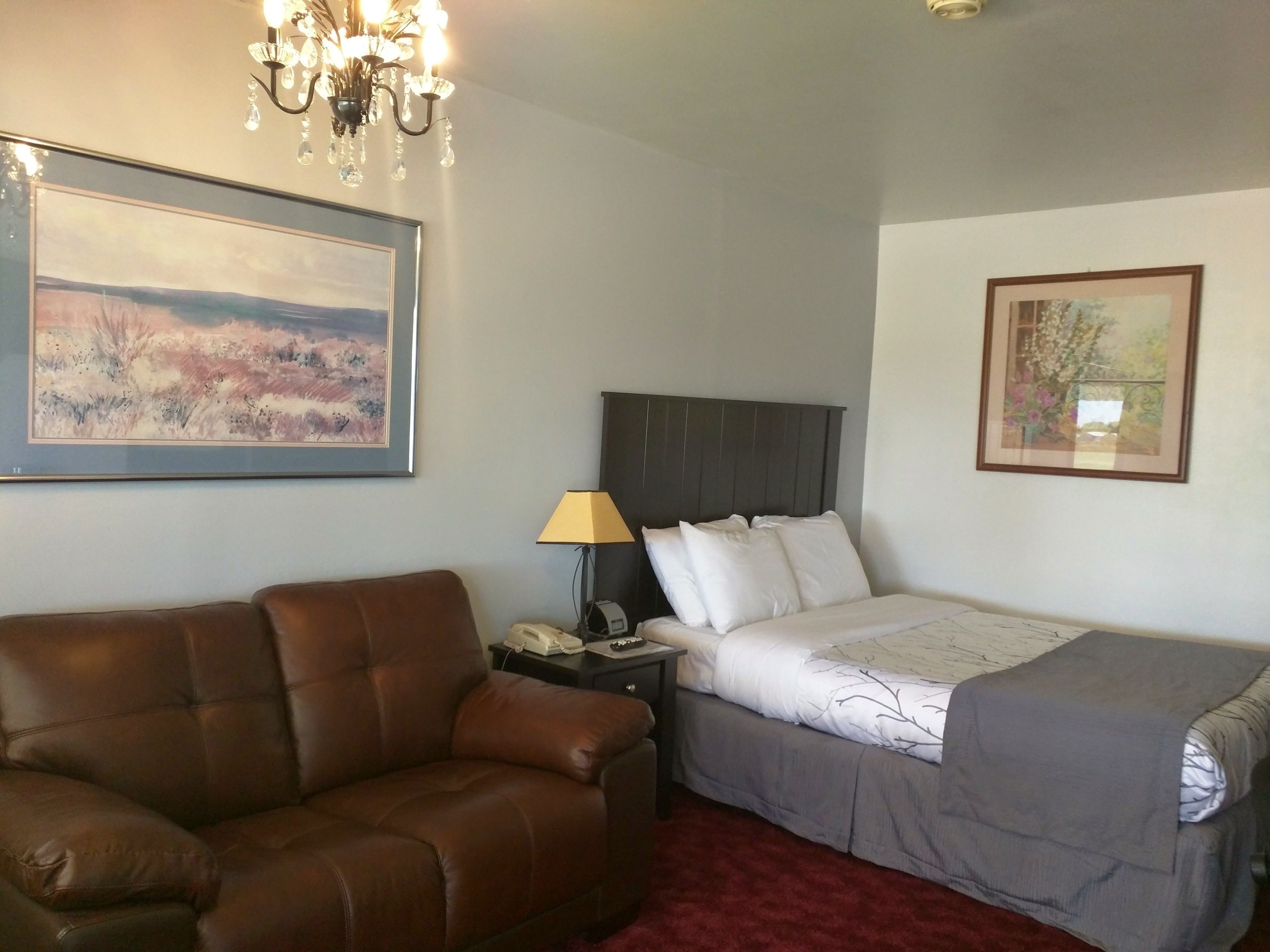 Habitación de hotel con un sofá de cuero marrón, una cama individual con una manta gris, una mesita de noche con una lámpara y dos cuadros enmarcados en las paredes.