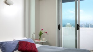 A bedroom with a bed, a small table with a vase of flowers, and a sliding glass door leading to a balcony with a city view. - Backpacker College at EC Vancouver: APT Living (Vancouver)