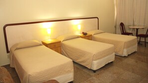 Habitación de hotel con tres camas individuales, cabeceros de madera, mesitas de noche con lámparas, una pequeña mesa redonda y una silla.