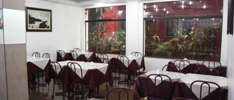A restaurant with multiple tables and chairs, large windows with plants, and framed artwork on the walls.