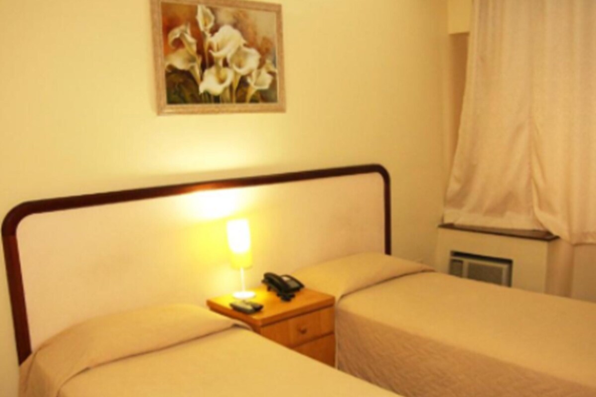 A hotel room with two beds, a bedside table with a lamp, a telephone, and a painting on the wall.