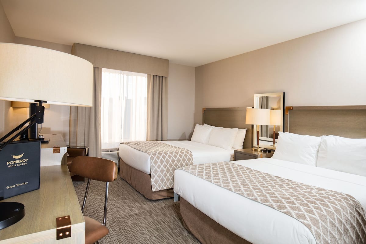 A hotel room with two beds, a desk, a chair, and a lamp.
