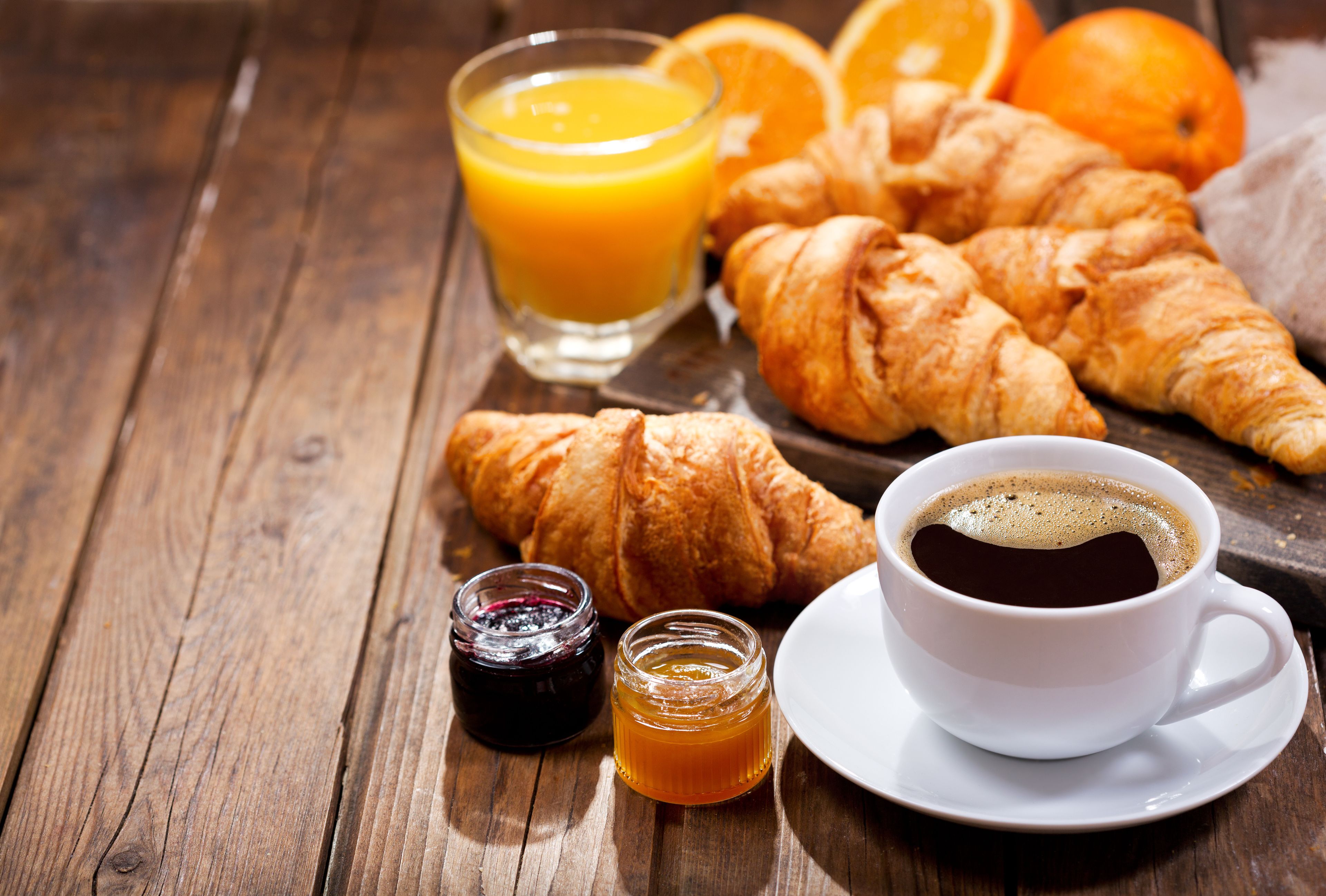 Een houten tafel met croissants, een glas sinaasappelsap, een kop koffie en twee kleine potjes jam.