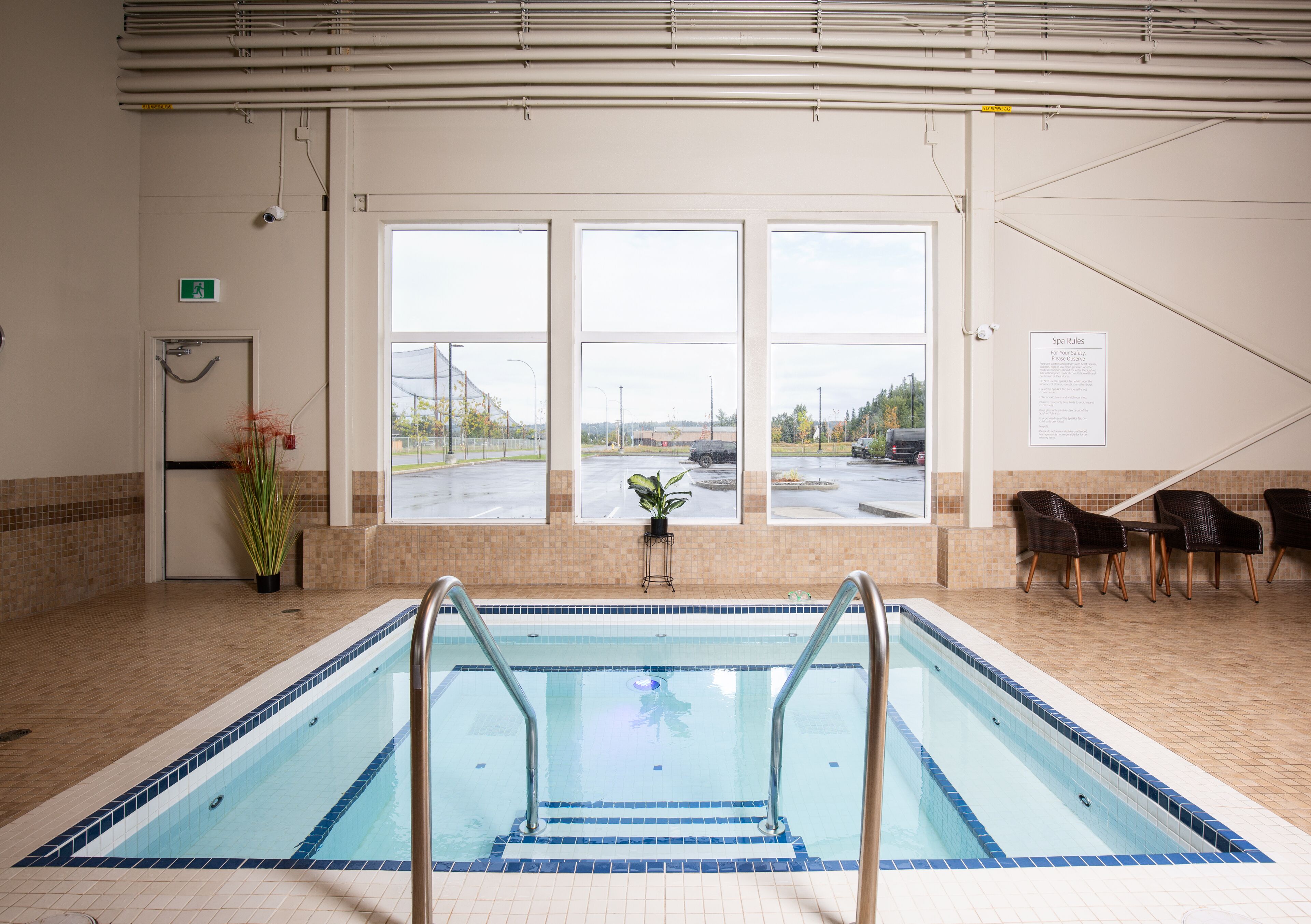 An indoor swimming pool with clear windows, a safety sign, and a seating area with chairs.