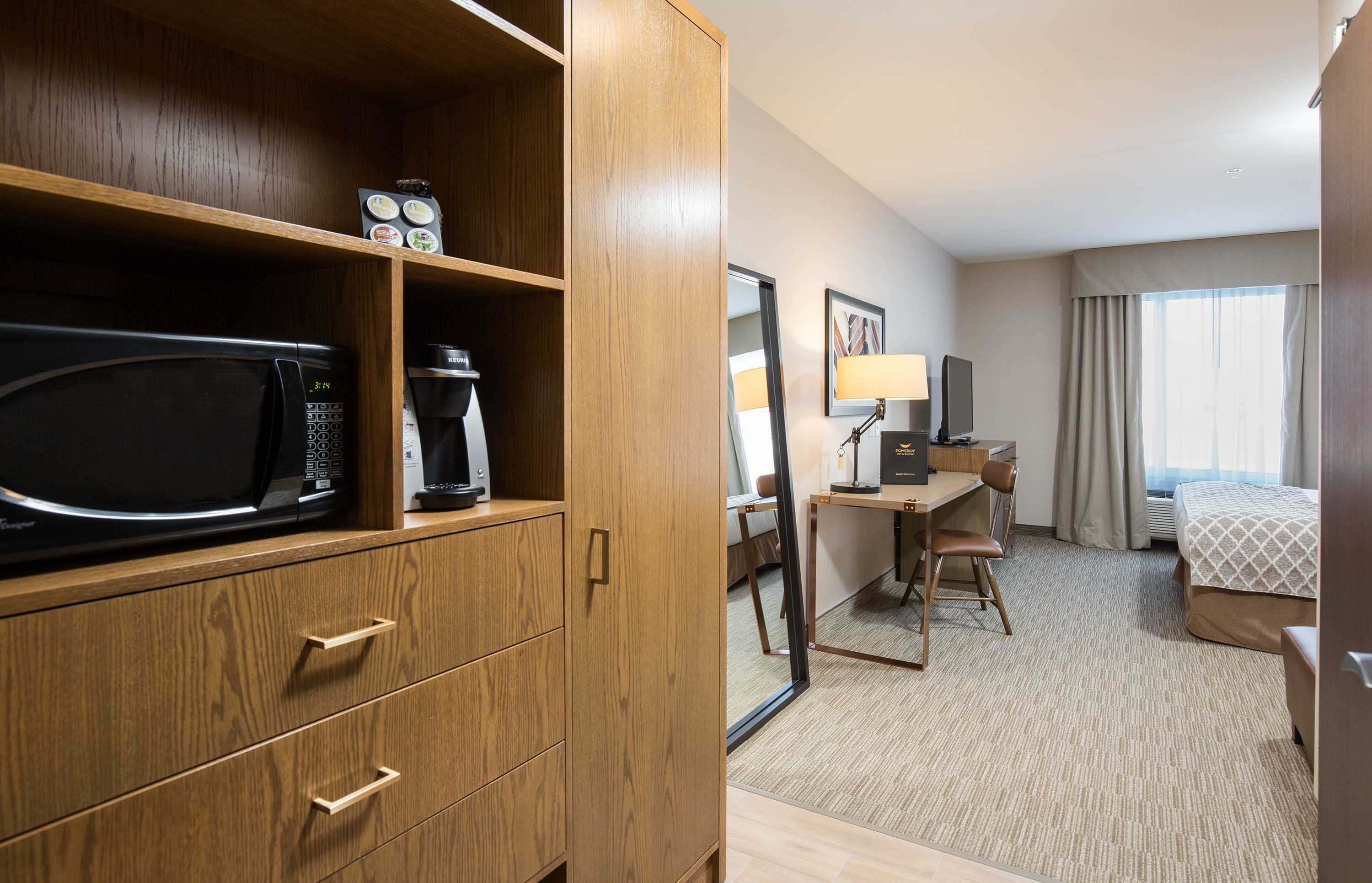 A hotel room with a wooden wardrobe, a microwave, a coffee maker, a desk with a chair, a bed, and a large window with curtains.