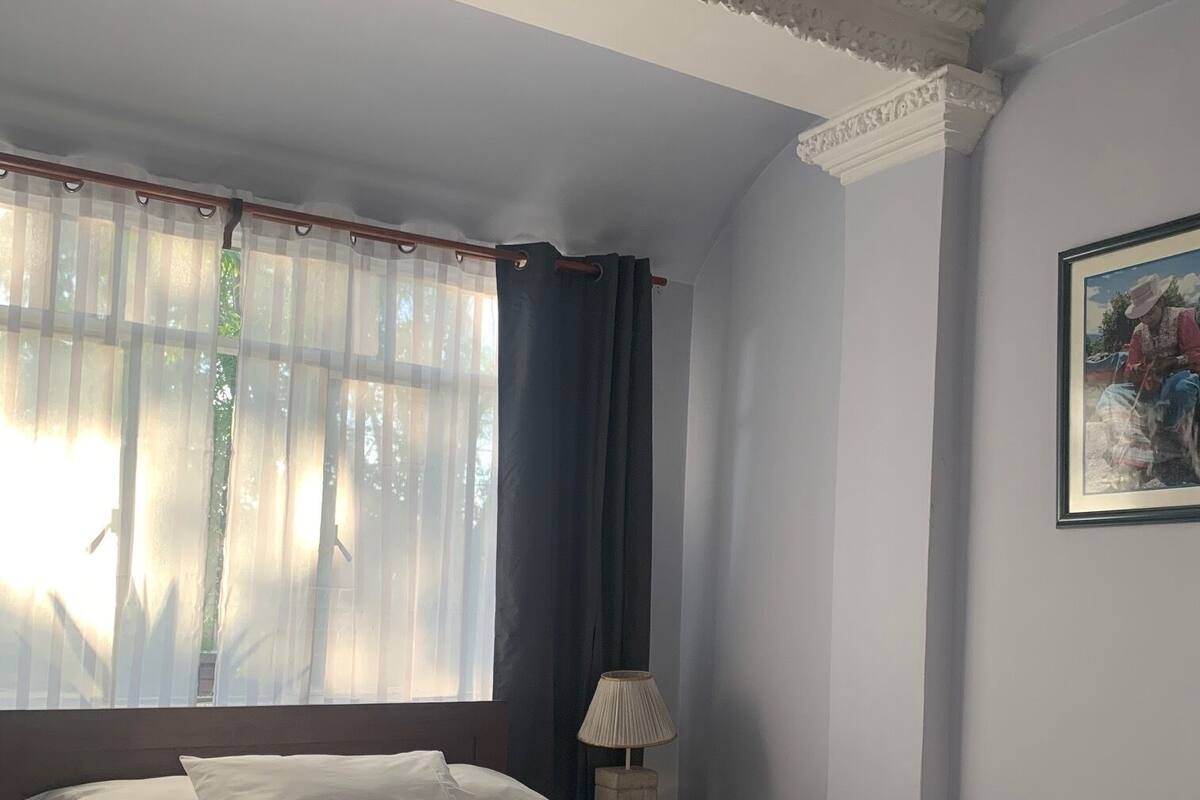 Ein Schlafzimmer mit Bett, Nachttisch mit Lampe, Fenster mit durchsichtigen Vorhängen und einem gerahmten Bild an der Wand.
