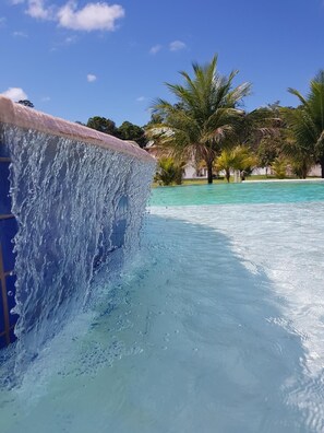 A clear blue pool with a waterfall feature and palm trees in the background. - RCR Green Field Casas para Temporada (Porto Seguro)