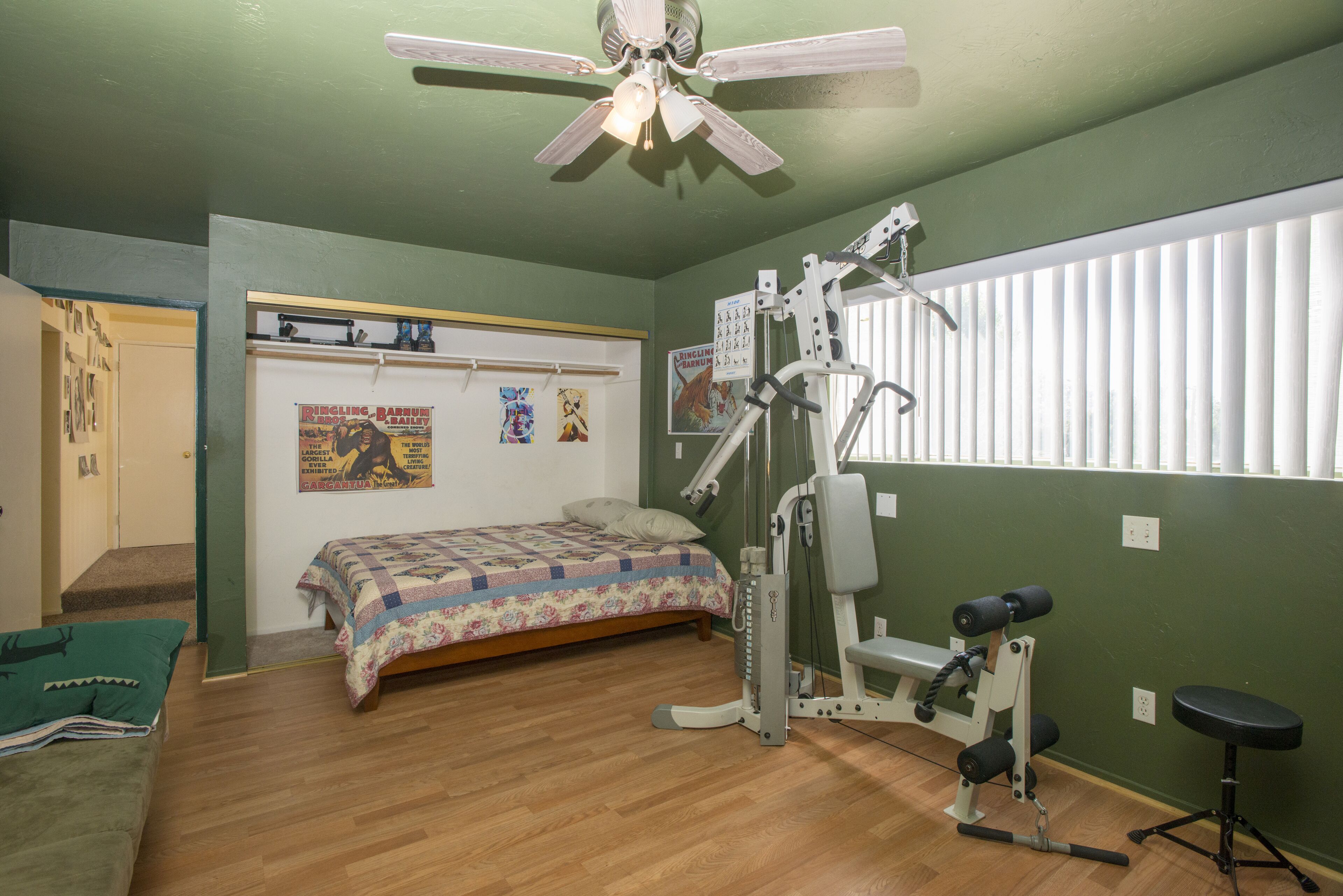 Une chambre à coucher comprenant un lit, du matériel d’exercice, un ventilateur de plafond et une fenêtre avec des stores.