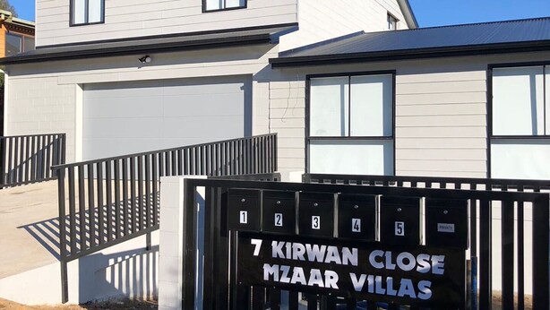 A modern villa complex with a gated entrance, numbered mailboxes, and a clear sky.