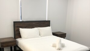 A neatly made bed with white linens and two pillows, a wooden headboard, and a small wooden nightstand with a drawer.