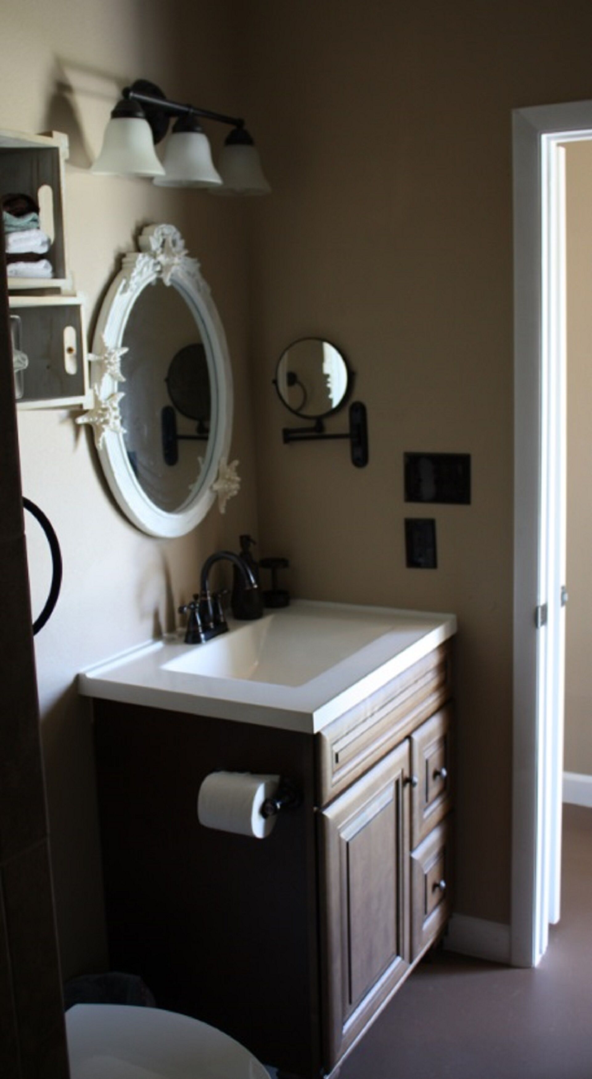 A bathroom with a white vanity, a round mirror, a wall-mounted light fixture, and a toilet paper holder.